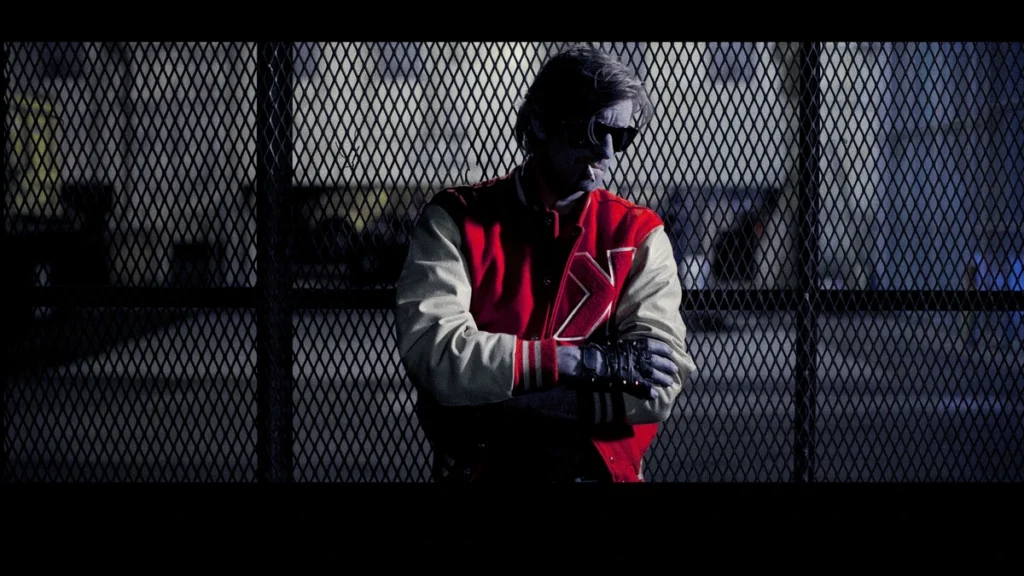 Kavinsky in a red and white varsity jacket behind a chain-link fence