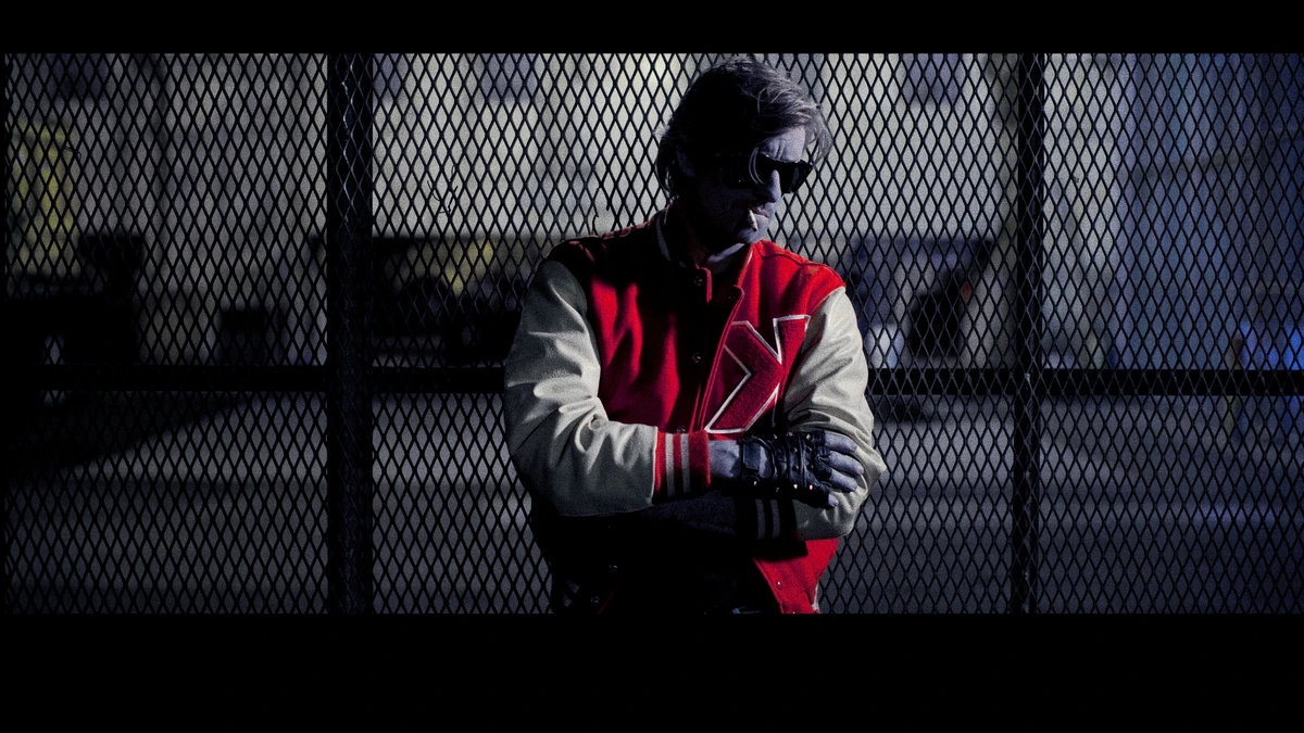 Kavinsky in a red and white varsity jacket behind a chain-link fence