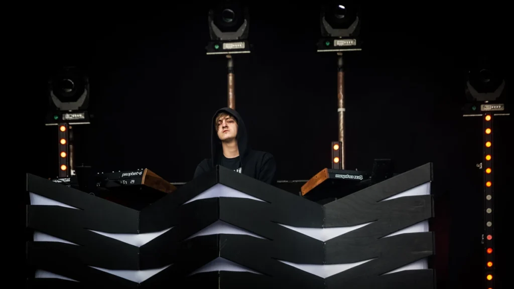 Perturbator performing behind a chevron-patterned booth under stage lights