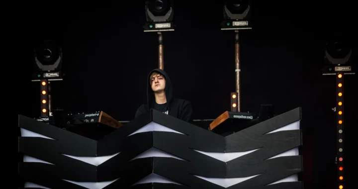 Perturbator performing behind a chevron-patterned booth under stage lights