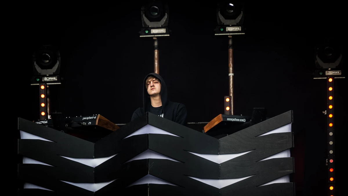 Perturbator performing behind a chevron-patterned booth under stage lights