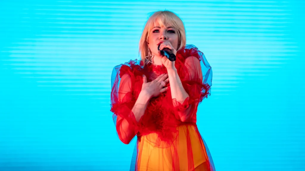 Carly Rae Jepsen singing into a microphone in a red dress against a bright cyan backdrop